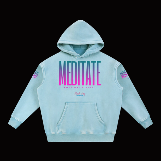 A light blue mineral-wash hoodie with a large 'MEDITATE' graphic in a pink-to-blue gradient on the chest, and smaller text reading 'BOTH DAY & NIGHT' below it. The sleeves also feature small pink and blue graphic details.